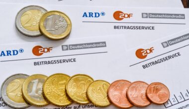 Auf mehreren Formularen des Beitragsservice für den Rundfunkbeitrag liegen Geldmünzen.