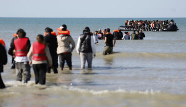 A shot of migrants wading through waters in life vests in the middle of the afternoon.