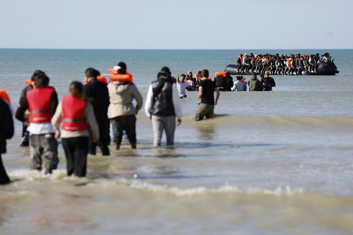 A shot of migrants wading through waters in life vests in the middle of the afternoon.