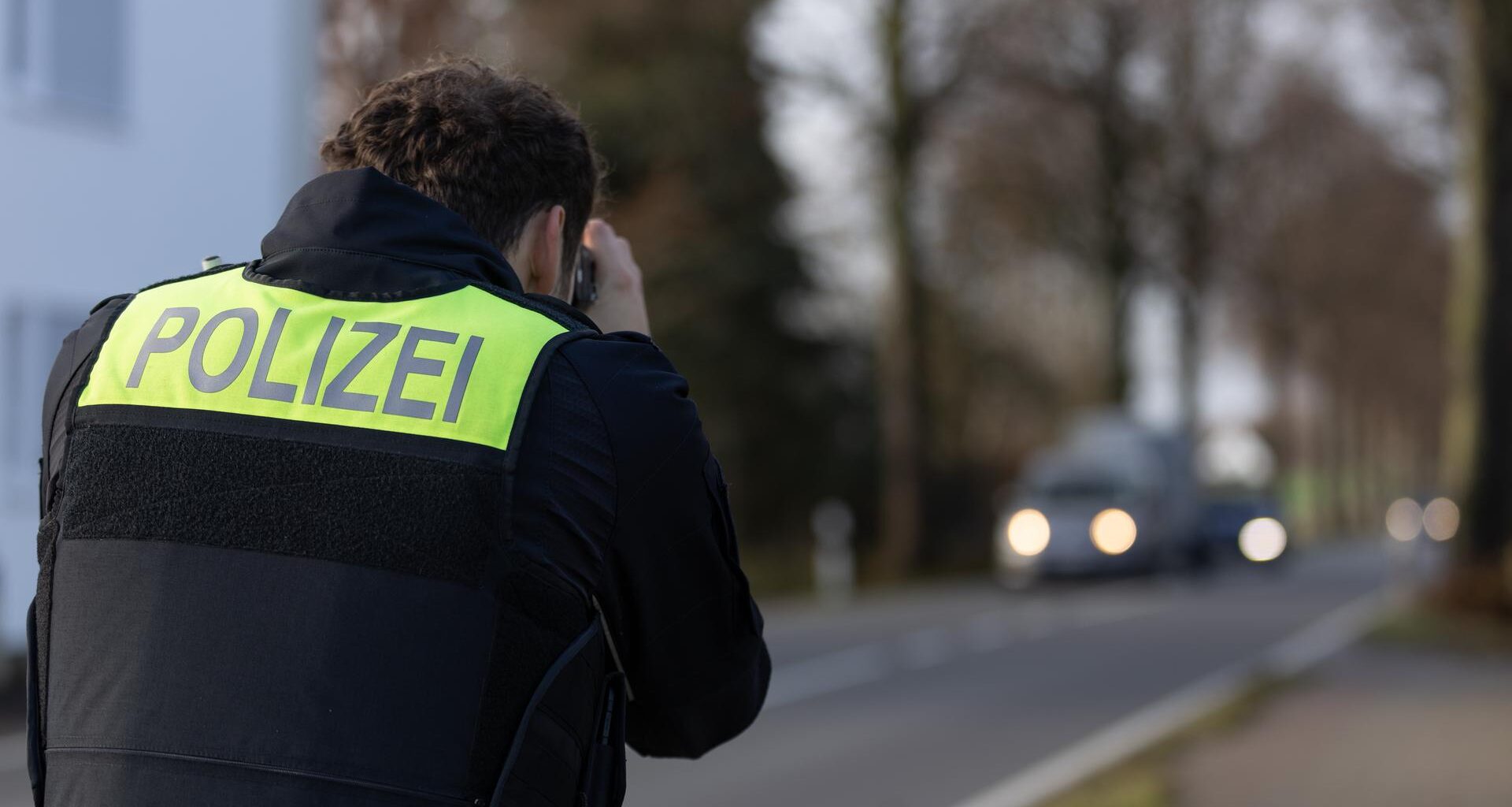 Ein Polizist zielt mit einem Laser für Geschwindigkeitsmessung auf heranfahrende Autos, man sieht ihn von hinten. Es ist düster, die Autos haben leuchtende Scheinwerfer.