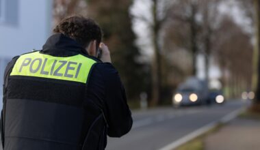 Ein Polizist zielt mit einem Laser für Geschwindigkeitsmessung auf heranfahrende Autos, man sieht ihn von hinten. Es ist düster, die Autos haben leuchtende Scheinwerfer.