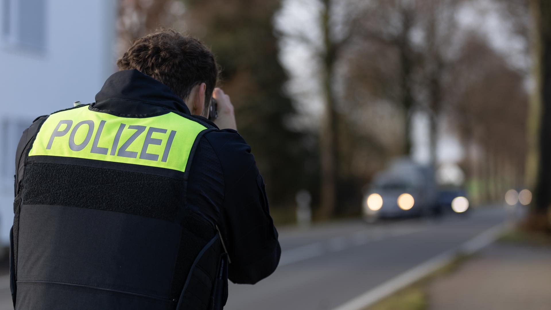 Ein Polizist zielt mit einem Laser für Geschwindigkeitsmessung auf heranfahrende Autos, man sieht ihn von hinten. Es ist düster, die Autos haben leuchtende Scheinwerfer. Ein Polizist zielt mit einem Laser für Geschwindigkeitsmessung auf heranfahrende Autos, man sieht ihn von hinten. Es ist düster, die Autos haben leuchtende Scheinwerfer.