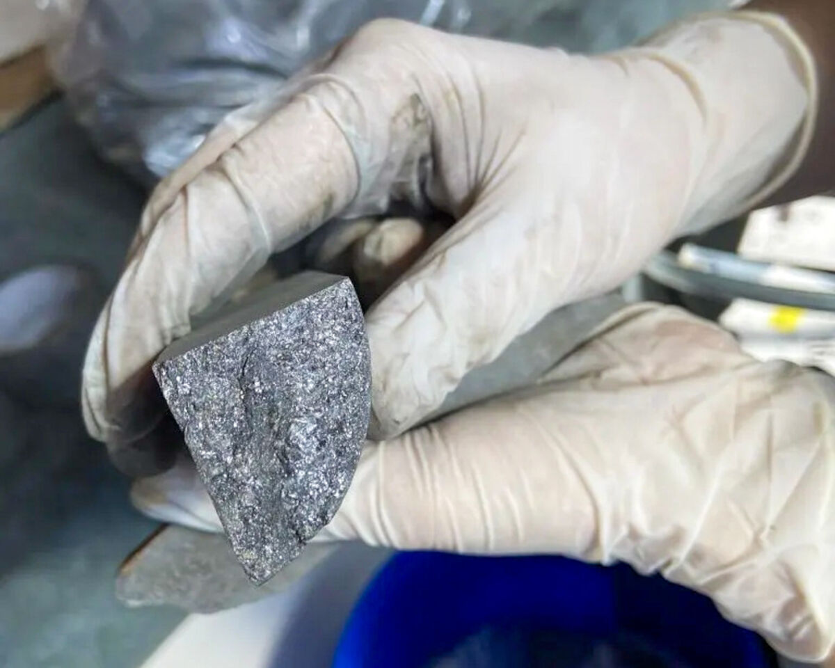 Gloved%20hands%20holding%20a%20shiny%20graphite%20sample%20above%20a%20blue%20bucket%2E