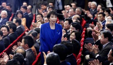 Takaichi in marineblauen Kostüm steht in den Parlamentsrängen zwischen den applaudierenden sitzenden Abgeordneten.