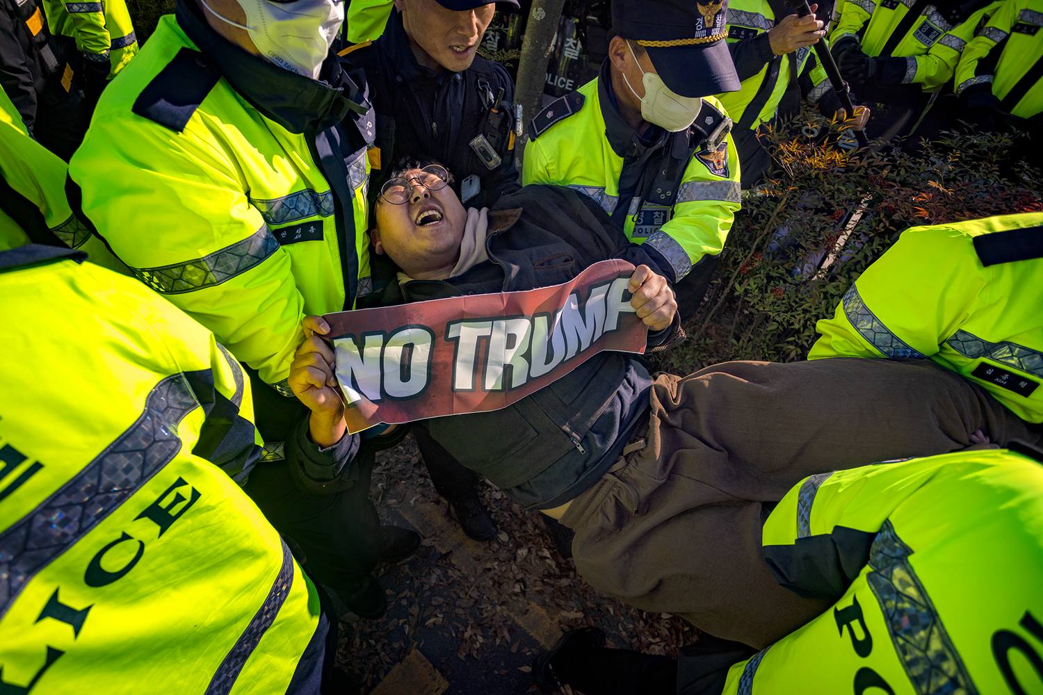 A man holding up a "NO TRUMP" sign is held by his arms and legs by police officers as they carry him away.