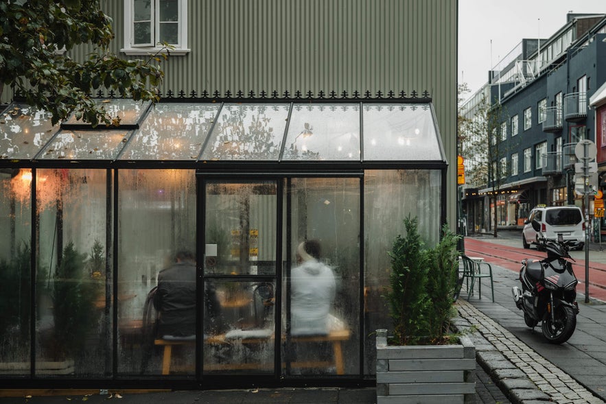 A cozy glass café in Reykjavik on a rainy day, with people sitting inside and a motorbike parked nearby on the wet street.