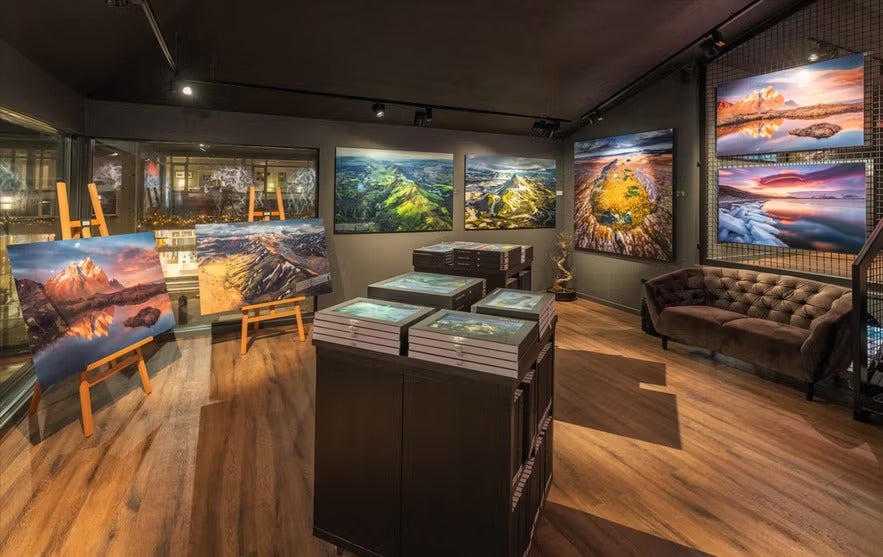An art gallery in Reykjavik displaying vibrant landscape photography of Iceland on walls and easels, with books on display and a brown sofa in the corner.