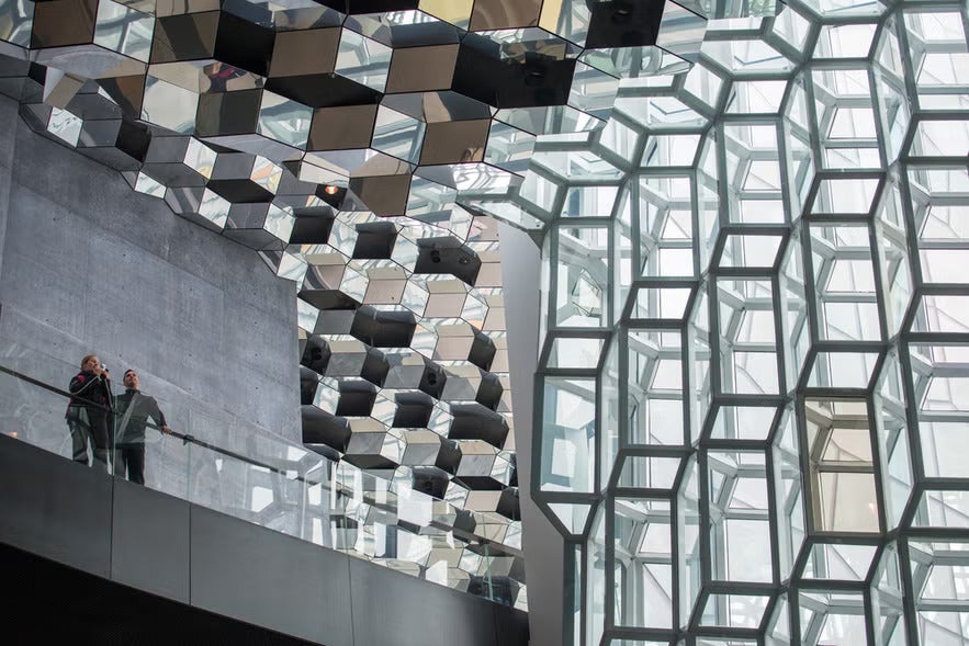 Interior view of Harpa Concert Hall in Reykjavik with its geometric glass facade and reflective ceiling, where two visitors stand on an upper walkway.