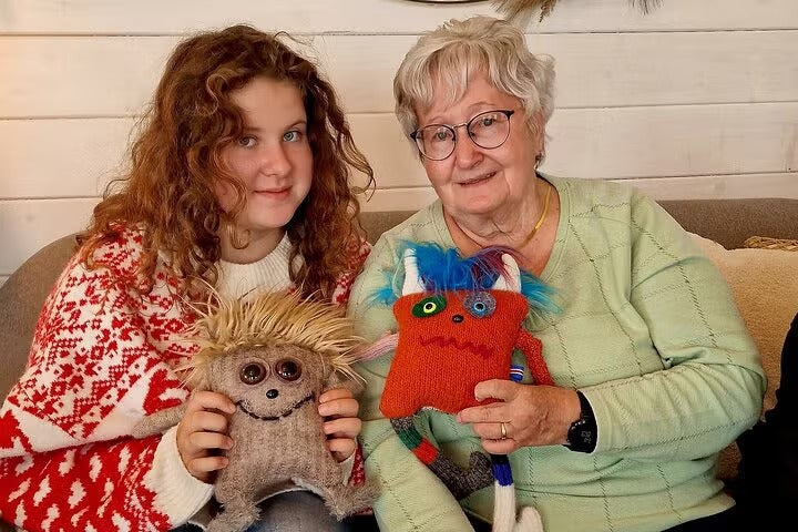 An elderly woman and a young girl sitting on a couch, smiling and holding colorful handmade knitted toys.