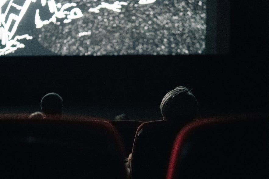 People watching a black-and-white movie in a dark cinema, viewed from behind red theater seats.