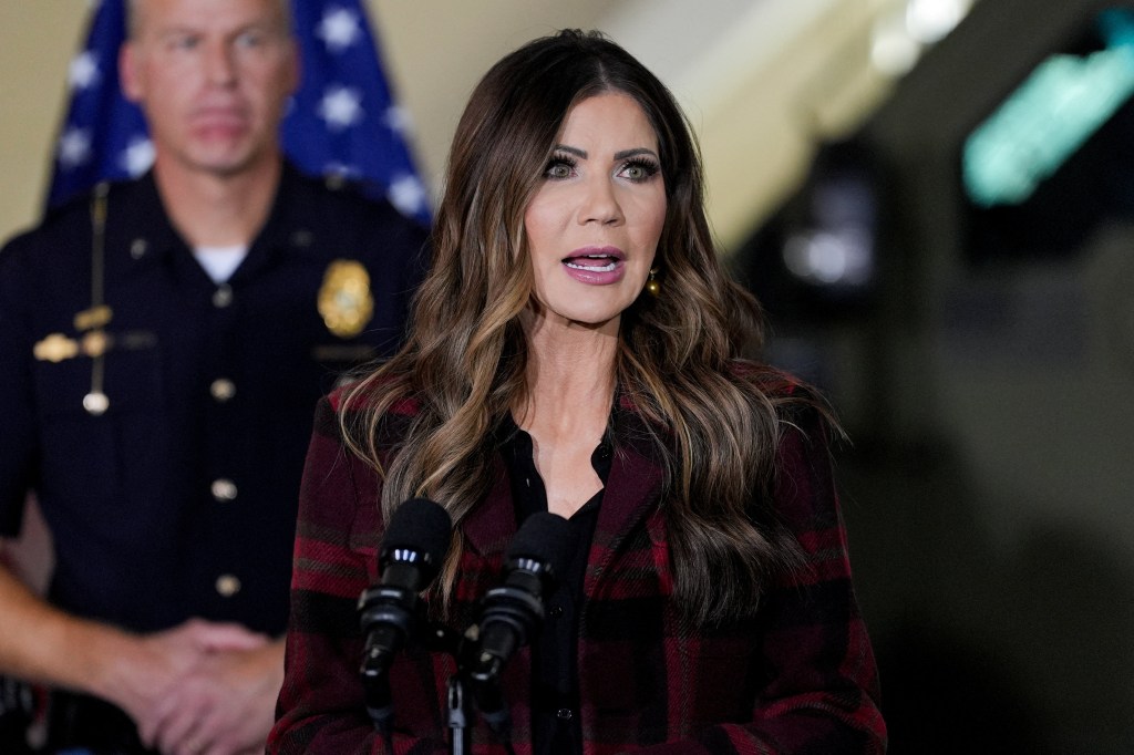 U.S. Homeland Security Secretary Kristi Noem speaks at a press conference, with a police officer standing behind her.
