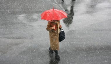 O femeie merge pe stradă cu o umbrelă roșie și se apără de lapoviță, iar pe jos este ud.
