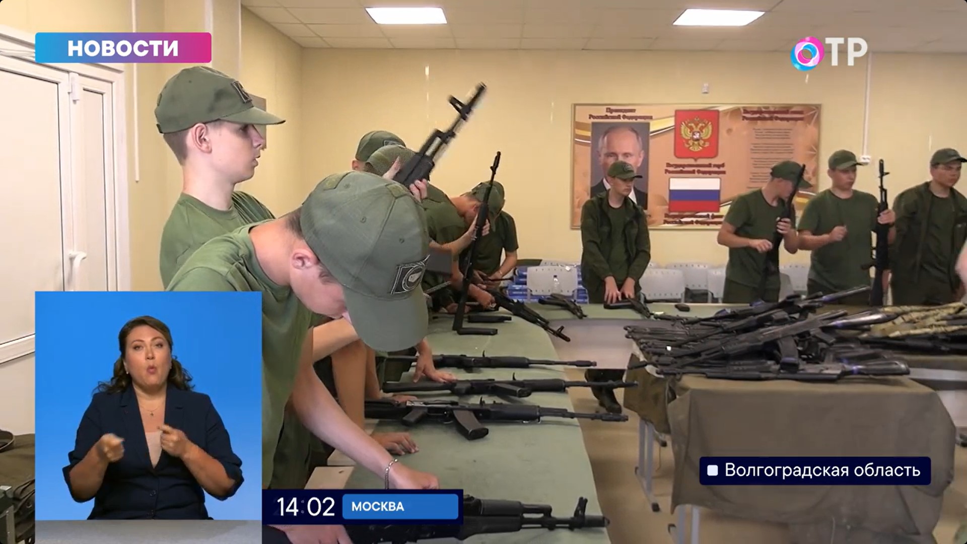 Teenagers in military-style uniforms assembling Kalashnikov assault rifles, with a television presenter in the lower-left corner.
