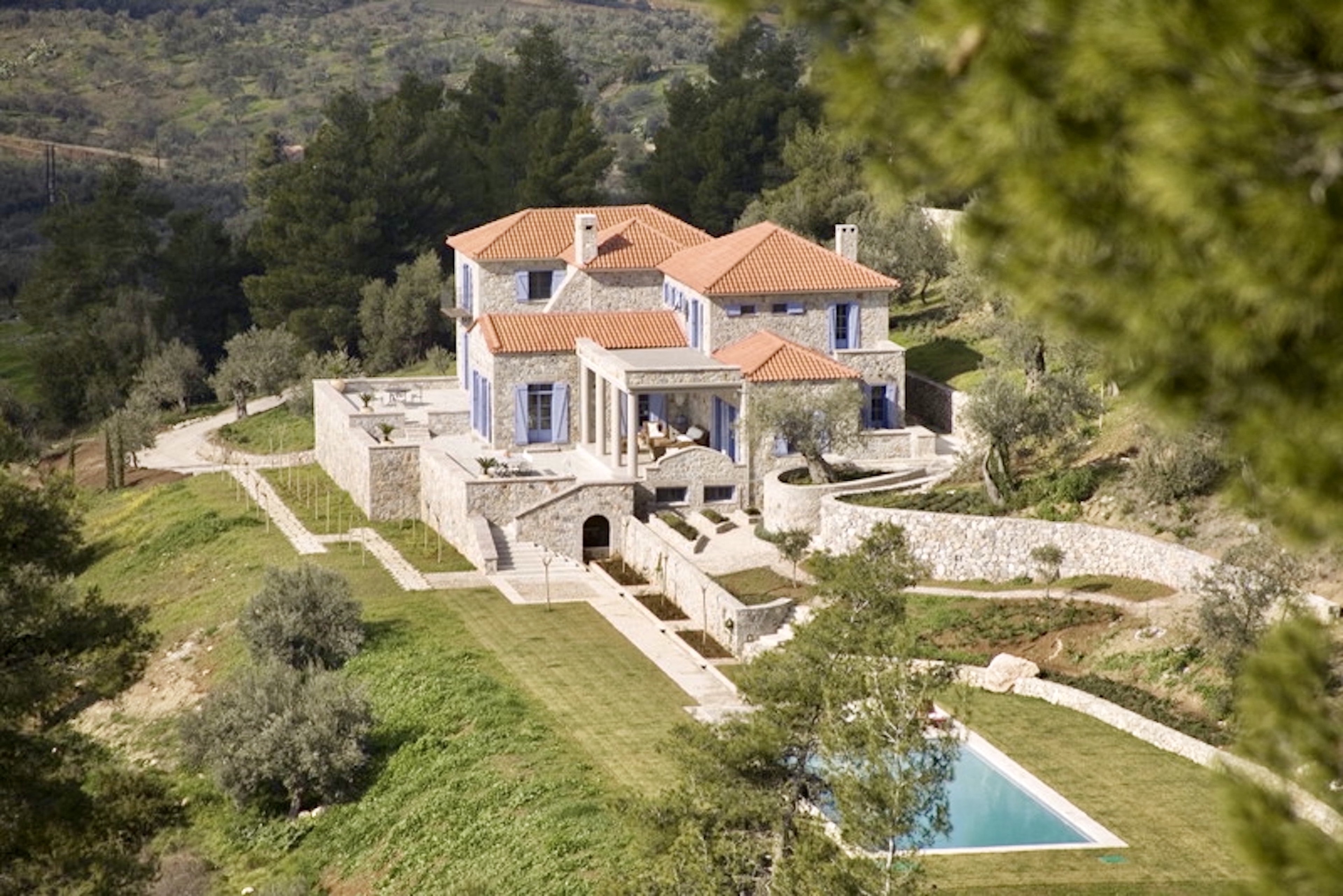 Vallea, a stone mansion with a red tile roof and blue shutters, sits on a hill with a swimming pool, surrounded by trees and terraces.