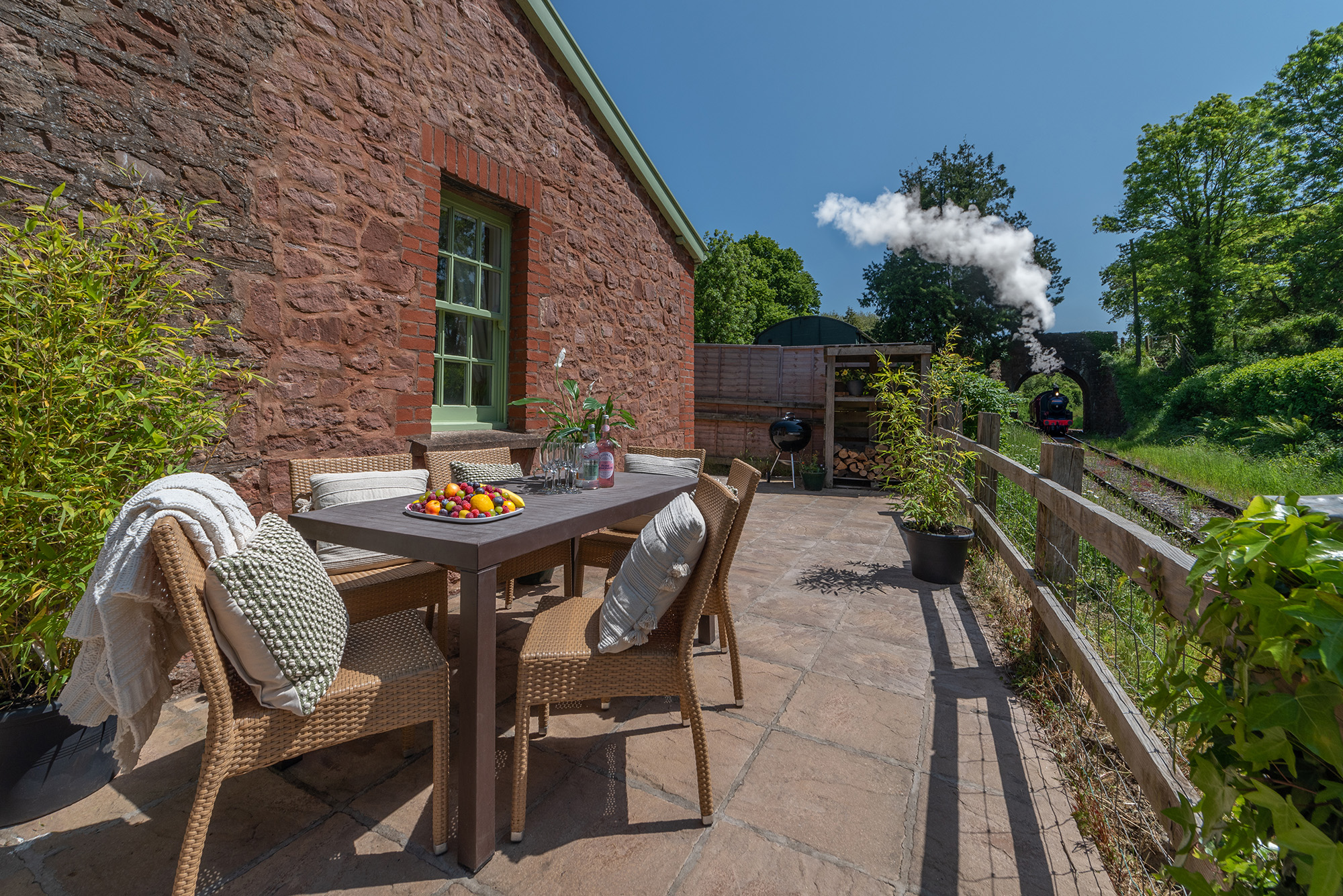 A patio table and chairs outside a brick cottage as a steam train emerges from a tunnel in the background.