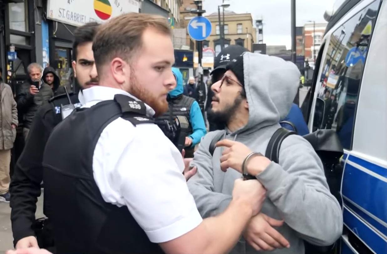 Police officer restraining a handcuffed man next to a police van.