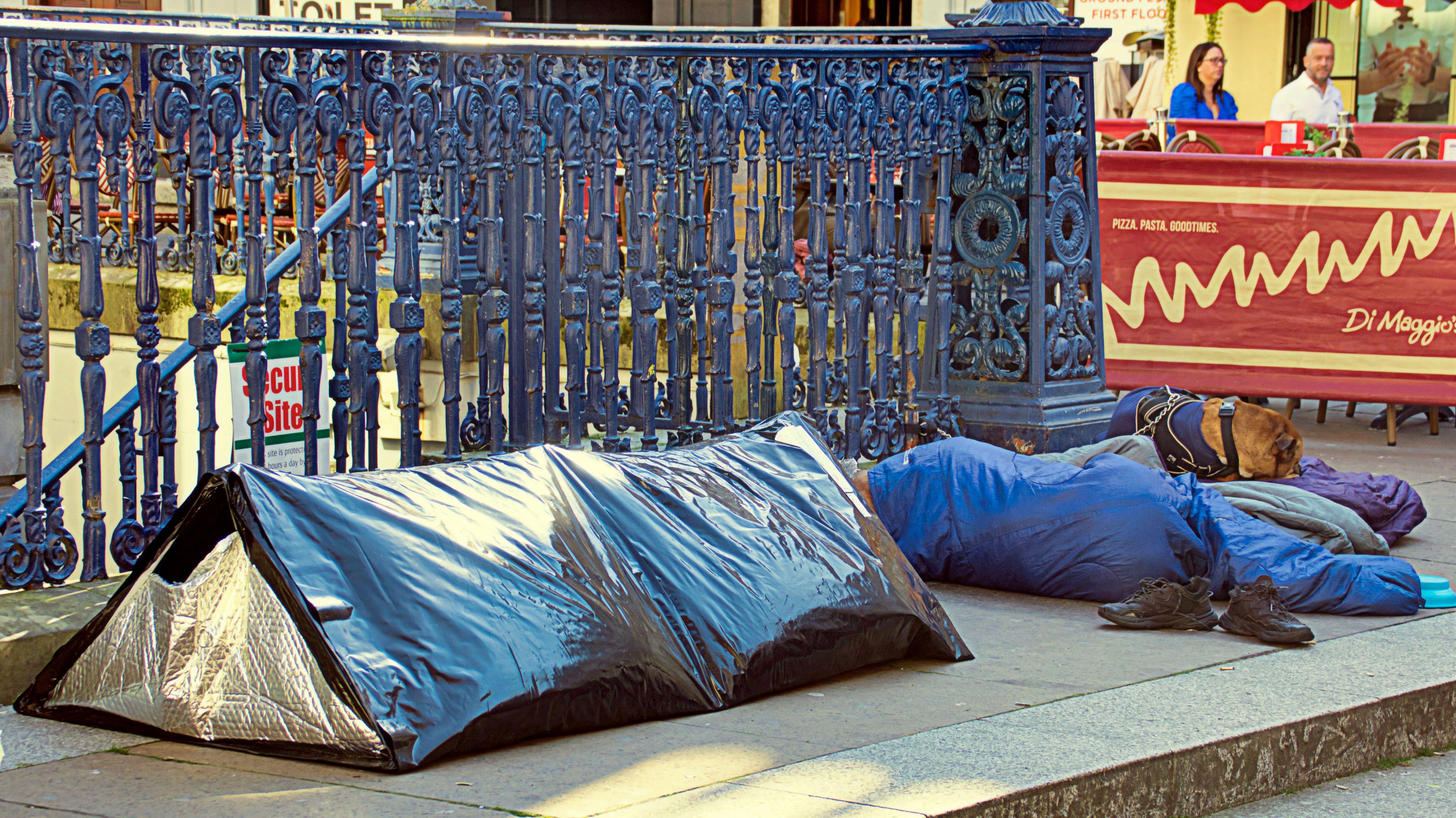 A person sleeping on the ground next to a small tent-like structure, with a dog beside them, near a metal railing and a restaurant sign.