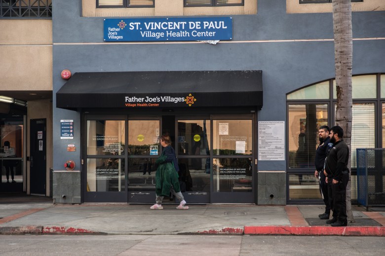 A person walks by the sliding doors of a building with a sign that says "St. Vincent De Paul Village Family Health Center" on it. Two security guards can be seen standing nearby.