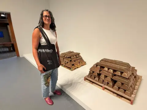 BBC / Regan Morris A woman wearing jeans and a tank top stands next to two piles of bronze ingots in a white display 