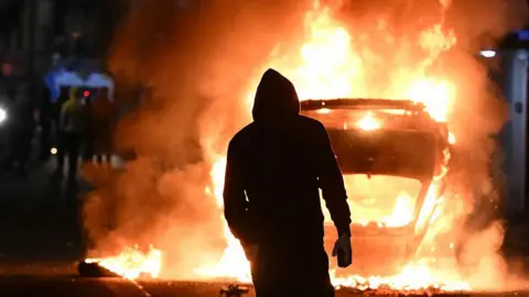 Getty Images A vehicle burns and a man stands silhouetted in front of it.