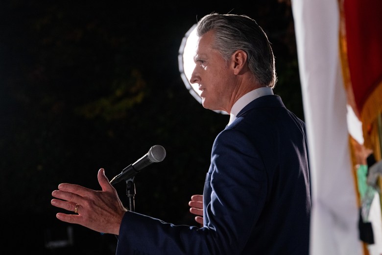 A side view of a person, wearing a blue suit, speaks while standing in front of a microphone. Another person, standing on the right and wearing a purple blazer, looks up towards the person in front of the microphone. An American flag can be seen nearby.