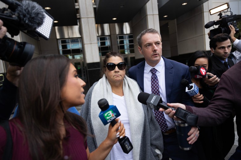 A person wearing sunglasses and a light shawl walks beside another person in a suit outside a courthouse, surrounded by reporters and TV crews holding microphones and cameras.