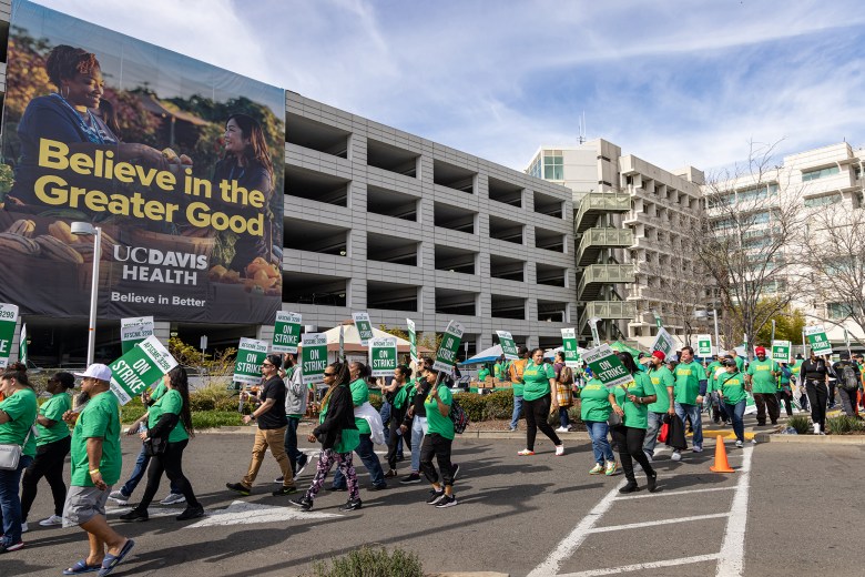 A wide view of a group of people in front of a building holding picket signs that read ‘AFSCME 3299 ON STRIKE,' near a large banner on the building that reads UC Davis Health.