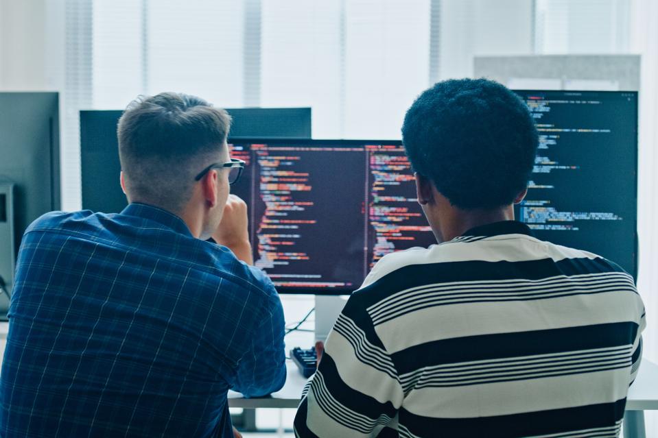 Two male software developers meeting on multiple computer screens displaying programming code, collaborating on project in office. Artificial intelligence and programming.