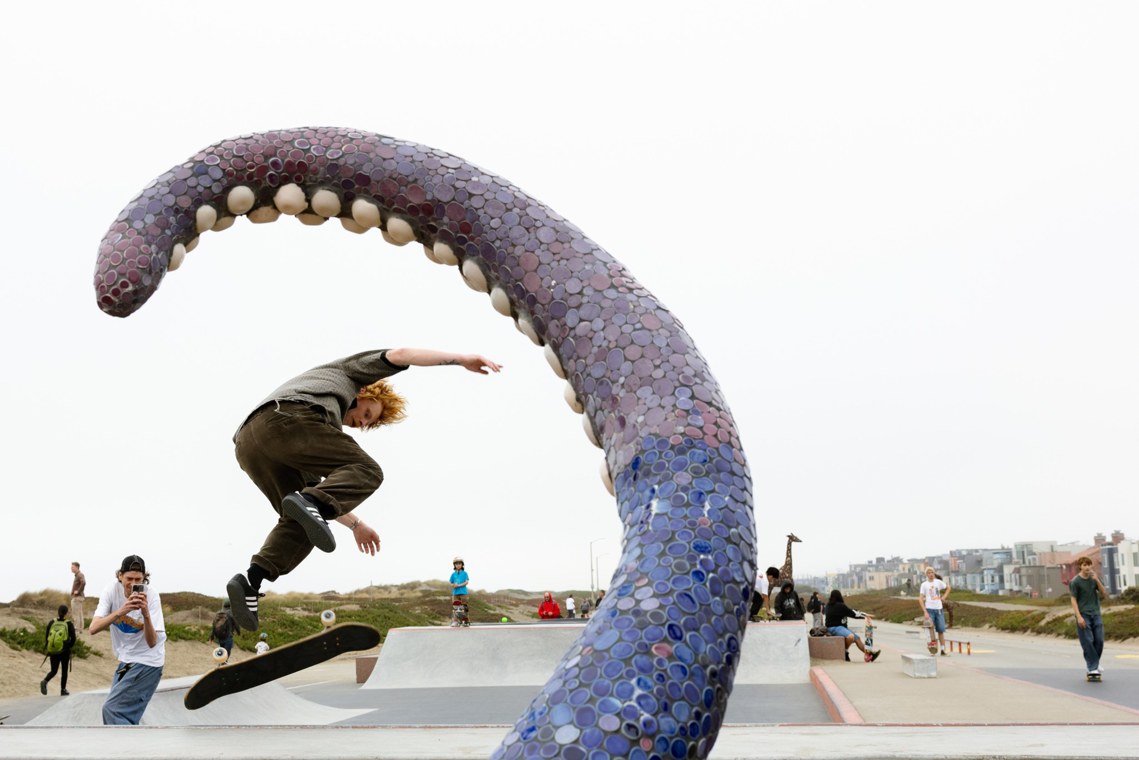 A skateboarder performs a trick mid-air near a large, curved purple tentacle sculpture at an outdoor skate park with people watching.