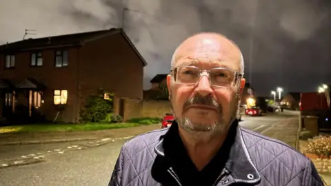 Andrew Turner/BBC Graham Plant, wearing glasses and sporting a beard and moustache, which is greying. He is wearing a black quilted jacket, and is standing under a street lamp in the evening on a housing estate.