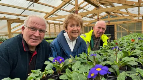 Andrew Turner/BBC Adrian Thompson, left, Joy Baldry and Glen Unstead, in a high visibility jacket, stand near rows of polyanthus in a timber-framed greenhouse. Mr Thompson and Mrs Baldry are wearing blue jackets.