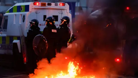 Reuters Armed police officers wearing a black uniform standing behind a white police jeep. There is flames rising into the air behind them.