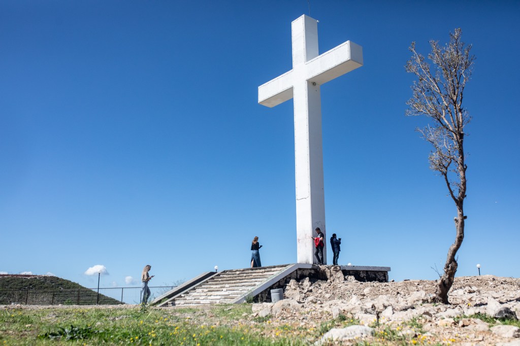 A large white cross on a hillside, with people standing near its base and a bare tree to its right, under a clear blue sky.