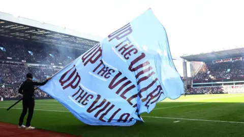 Reuters A blue Aston Villa flag is waved inside the stadium before a match. It says Up the Villa on it in maroon writing. Fans can be seen in grandstands around the pitch.