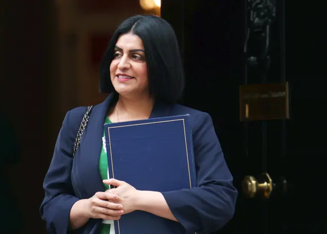 Shabana Mahmood in a blue blazer, holding a blue folder. Stood in front of a black door.