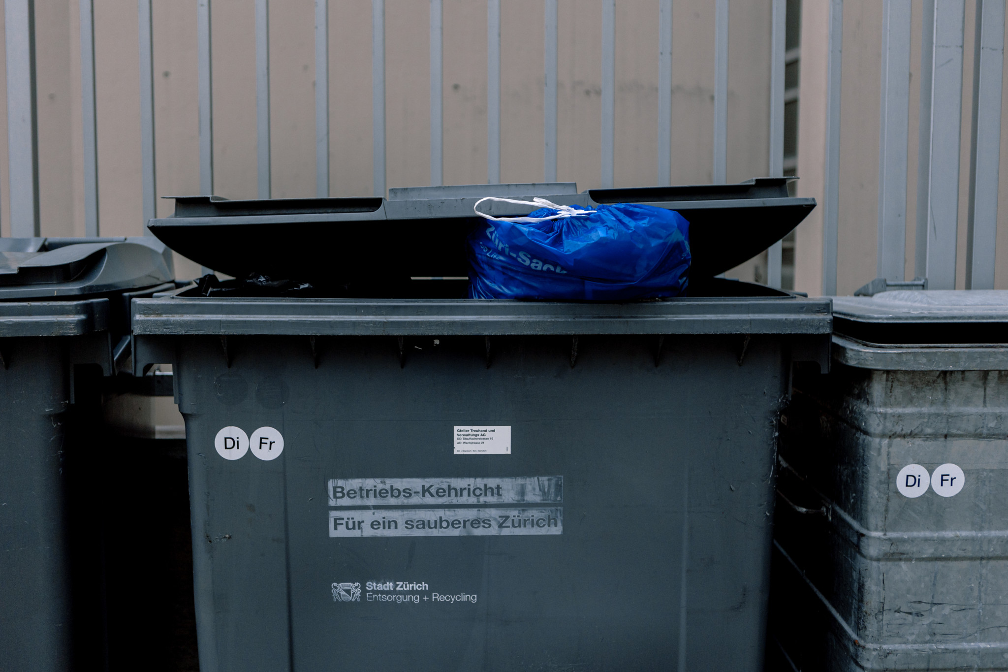 Ein blauer Züri-Sack liegt auf einem geöffneten grauen Müllcontainer in Zürich. Der Container ist mit Aufklebern für Müllabholungstage und einem städtischen Slogan versehen.