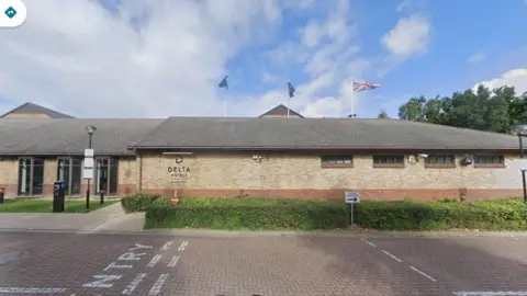 Google Single-storey brick-built hotel with small brown-framed windows and a grey pitched roof. There are entrance doors set back on the left. There is a low hedge in front of the building and a roadway in the foreground of the picture. There are trees in the background to the right.