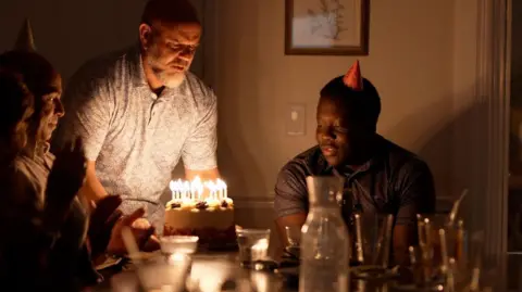 Liam Weir/BBC Marven looking on and smiling as a man brings a birthday cake with lit candles to the table where he and some other people are sitting. The lights are low and Marven is wearing a party hat.