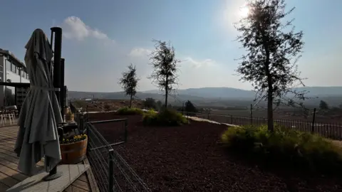 The view from Amichai's winery. There is a deck with an umbrella folded. As you look our from it there are three trees, then a fence then field and hills in the background. There is blue sky with a hint of cloud and the sunlight gives the image a misty feel