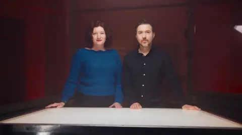 Tom Barnes/Channel 4 Turi King, wearing a blue long-sleeved blouse, and Alex Kay, wearing a black shirt, standing at a desk.