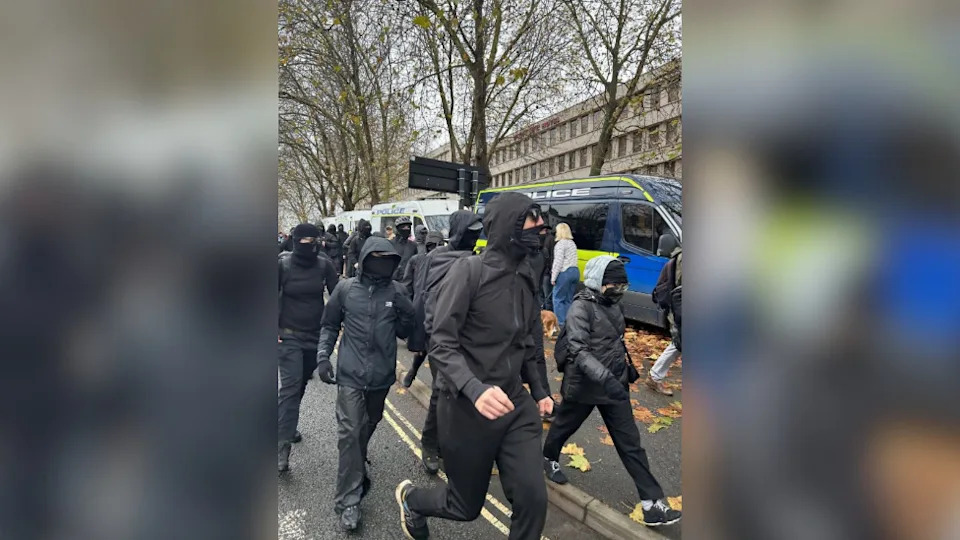 A group of people dressed all in black with black balaclavas on a city street. A line of police vans are visible in the background. A large grey building and a row of street trees are also visible.