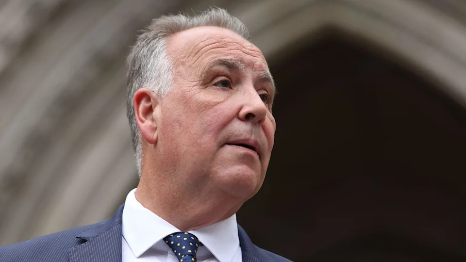 Chris Whitbread is dressed in a formal dark blue suit paired with a white shirt and a navy tie featuring a pattern of small yellow dots. He is standing in front of point archways belonging to the High Court in London.