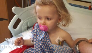 Young girl in a hospital bed wearing a blue bed-shirt. She looks poorly and has a pink dummy in her mouth.