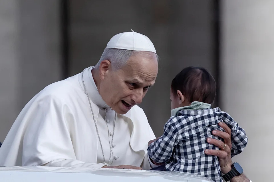 A man in religious attire gently holds and looks at a baby wearing a checkered shirt, creating a moment of connection and tenderness