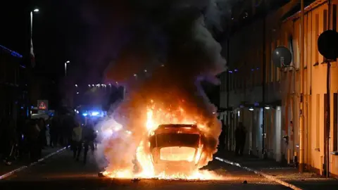Getty Images A car lying on its roof in the middle of a road. It is on fire. There are houses on either side of the street.
