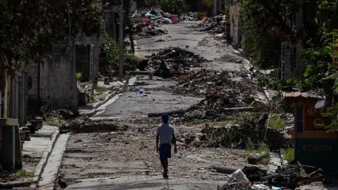 CLARENS SIFFROY/AFP via Getty Images A wide shot of a lone figure walking along a road covered in debris in the Delmas 30 neighborhood in September 2025