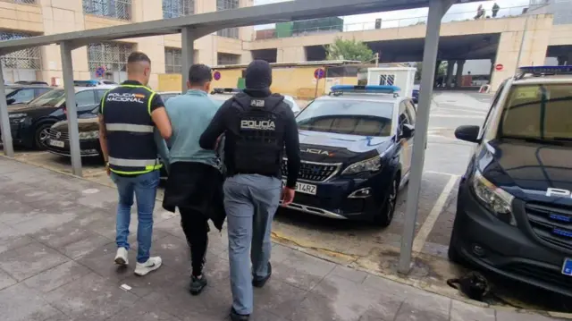 Un hombre con camiseta azul de manga larga flanqueado por dos agentes de Policía protegidos por chalecos balísticos. Caminan hacia autos policiales estacionados. 