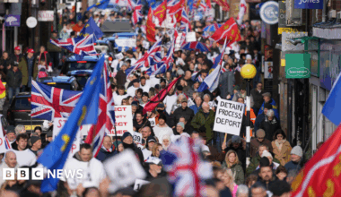 Hundreds of people marching on a town high street. Some people are carrying union jacks and Sussex county flags, while one person holds a sign that reads "detain process deport".