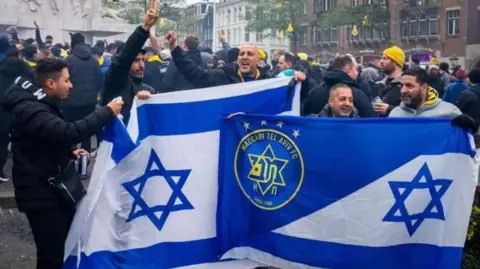 AFP via Getty Images Fans of Maccabi-Tel Aviv hold club flags as they sing out in support of their club, as other fans are gathered behind them, in an Amsterdam square.
