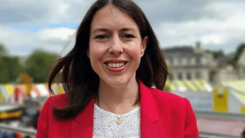 MP Alice Macdonald in front of Norwich Market. She is wearing a red jacket over a white top. She has shoulder-length dark hair and is smiling at the camera. The colourful striped roofs of the market stalls are behind her. 